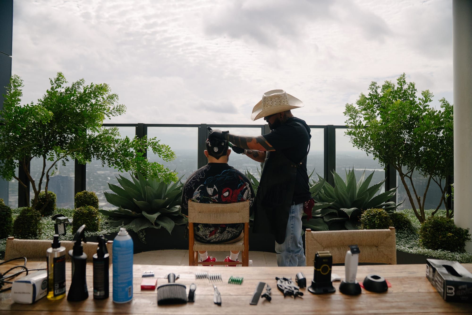 Roy cutting a client's hair on a rooftop above the city, his clippers and combs laid out on a table in front